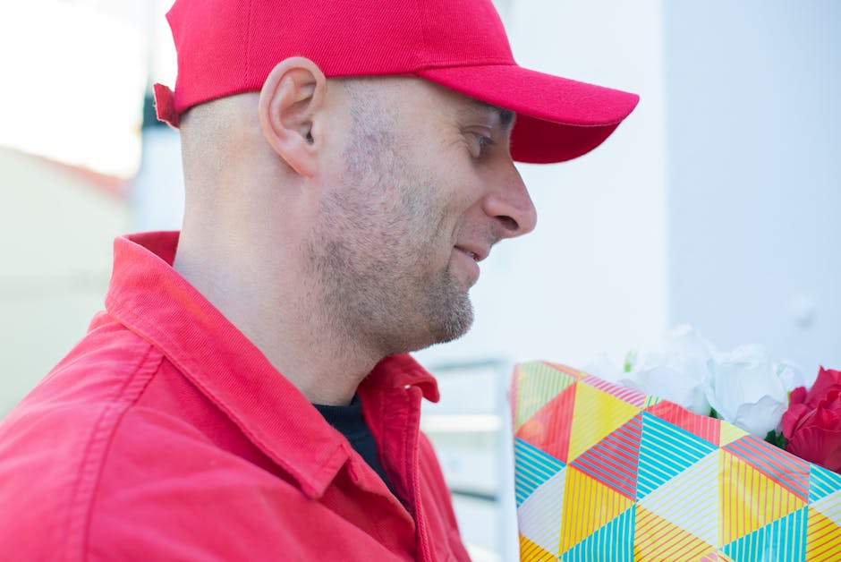 A delivery person wearing a red cap and matching red jacket is holding a bouquet of fresh flowers wrapped in striped, colorful paper, with pink roses and white blossoms visible. The scene is set outdo