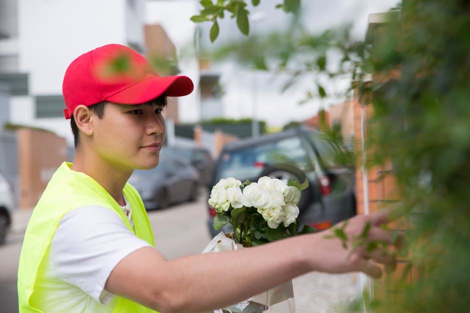 A young delivery person wearing a red cap and a neon yellow vest is holding a bouquet of fresh white hydrangeas wrapped in white paper, preparing to place the floral gift on a doorstep. The arrangemen