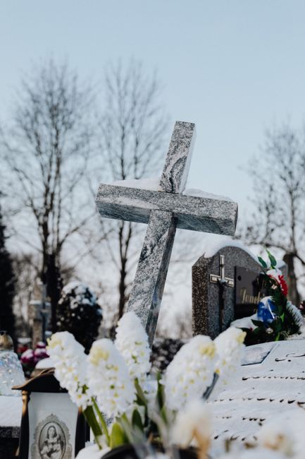 A close-up of a large grey granite cross gravestone set amidst a snowy cemetery landscape with leafless trees in the background under a clear sky. At the base of the cross, there are white hyacinth fl