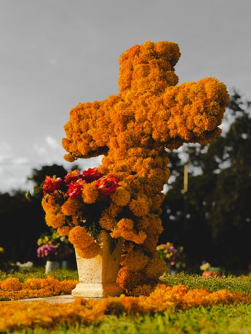 A floral cross composed of vibrant orange marigolds and other orange flowers, assembled as a grave memorial, housed in a stone pot on a pedestal. The arrangement features a standing cross shape, with 