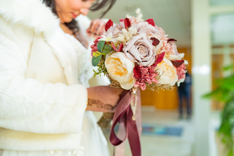 A person holding a floral bouquet with a mix of large, soft pink and cream roses, deep red and pink flowers, and green foliage, wrapped with a dark pink ribbon, in an indoor setting with natural light