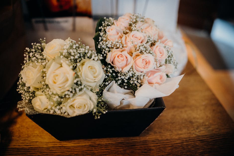 A black rectangular box containing two bouquet arrangements of fresh roses on a wooden surface. One bouquet features white roses accented with small white baby's breath flowers, while the other showca