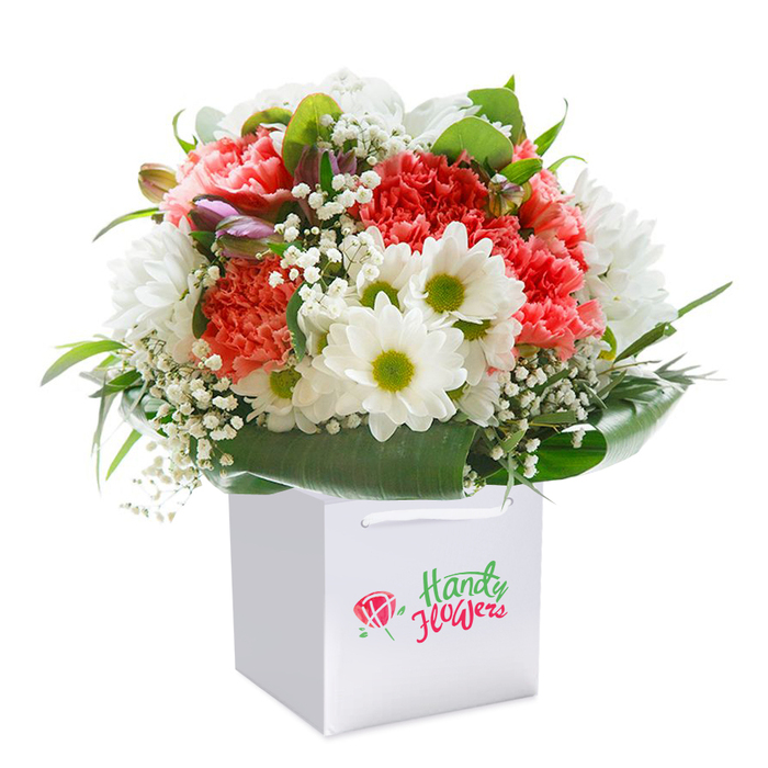 Standing tall in a crisp white square gift bag, this exuberant bouquet is like a burst of happiness delivered straight from the heart of Chase Cross. Its centrepieces-voluminous coral, salmon, and deep red carnations-unfurl their ruffled petals in profusion, offering a soft, inviting feel and a lively play of colour. Scattered among them are immaculate white daisies, their bold lime-yellow centres sparkling like morning sunlight over nearby Clockhouse Gardens. The bouquet is touched with feathery baby's breath, lending an ethereal airiness and subtle innocence to the composition. Subtle purple-tipped closed buds quietly enrich the texture at the edge, adding a whisper of curiosity and future colour. Embracing everything are lush, glossy green leaves that arc around the base, drawing the eye and providing a natural, supportive nest for the flowers. The arrangement fills its gift bag with a rounded, abundant silhouette, signifying both skill and a wish to delight. The bag's neat ribbon handles stand ready for easy transport, ideal for doorstep delivery or in-person gifting around Chase Cross. Perfectly lit, the image lets each hue and texture shine-a celebration of the tactile craft at the hands of local florists who take pride in making every occasion special, from birthdays and anniversaries to simple, thoughtful gestures. This bouquet captures the vibrant yet grounded essence of the Chase Cross community in floral form.