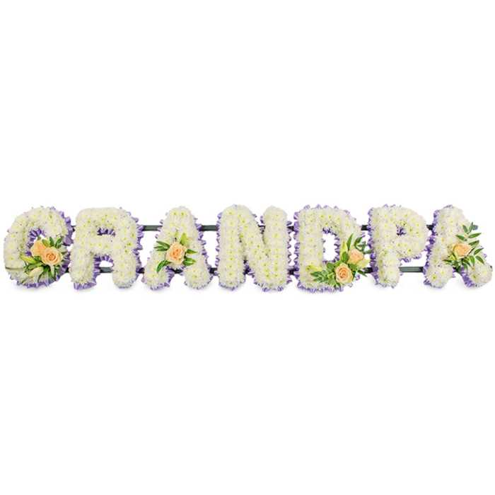The photograph showcases a bespoke funeral flower tribute spelling the word "GRANDPA" in tall, block capital letters, aligned in a continuous horizontal row as if resting on a stand at a memorial in Chase Cross. Each letter is expertly coated in dense clusters of fresh white chrysanthemums, or similar small daisy-like blooms, giving the surface a rich, textured appearance. The tiny petals sit close together, creating a soft yet structured look that feels both comforting and dignified. Around the outside of every letter runs a fine edging of light lavender or soft lilac flowers, which carefully traces the contours of the G, R, A, N, D, P, and A. This subtle purple border provides visual separation and depth, gently highlighting the shapes without overwhelming the serenity of the white base. Interwoven into the design are small clusters of accent flowers that add warmth and individuality. The initial 'G' features a spray of peach roses-three or four blooms at different stages of opening-offset by slender white lily buds and glossy green foliage that spills slightly over the white flowers. The first 'A' and the last 'A' each host a single, delicately placed peach rose, surrounded by fresh greenery, suggesting simple yet heartfelt tributes from family. The 'D' repeats the composition of the 'G' with another compact rose cluster. These peach accents bring a gentle glow to the arrangement, evoking fond memories and affection. A slender dark bar or frame supports the letters underneath, giving the tribute stability for display at local churches, chapels, or cemeteries near Chase Cross. Photographed against a bright white backdrop, the composition appears clear and uncluttered, conveying quiet sorrow, gratitude, and love for a cherished grandfather.